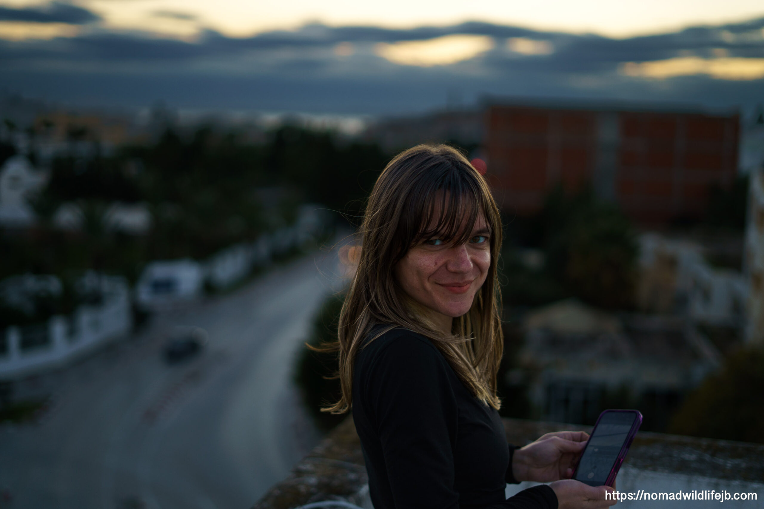 Woman standing on a rooftop in Hammamet at dusk, looking back toward the camera with the city blurred behind her