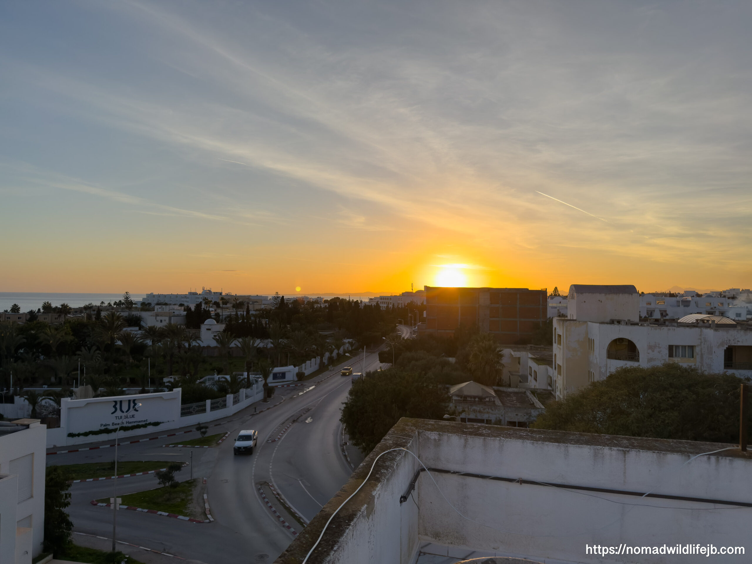 Sunset over a coastal city with roads, buildings, and palm trees visible, viewed from above as the sun dips toward the horizon.