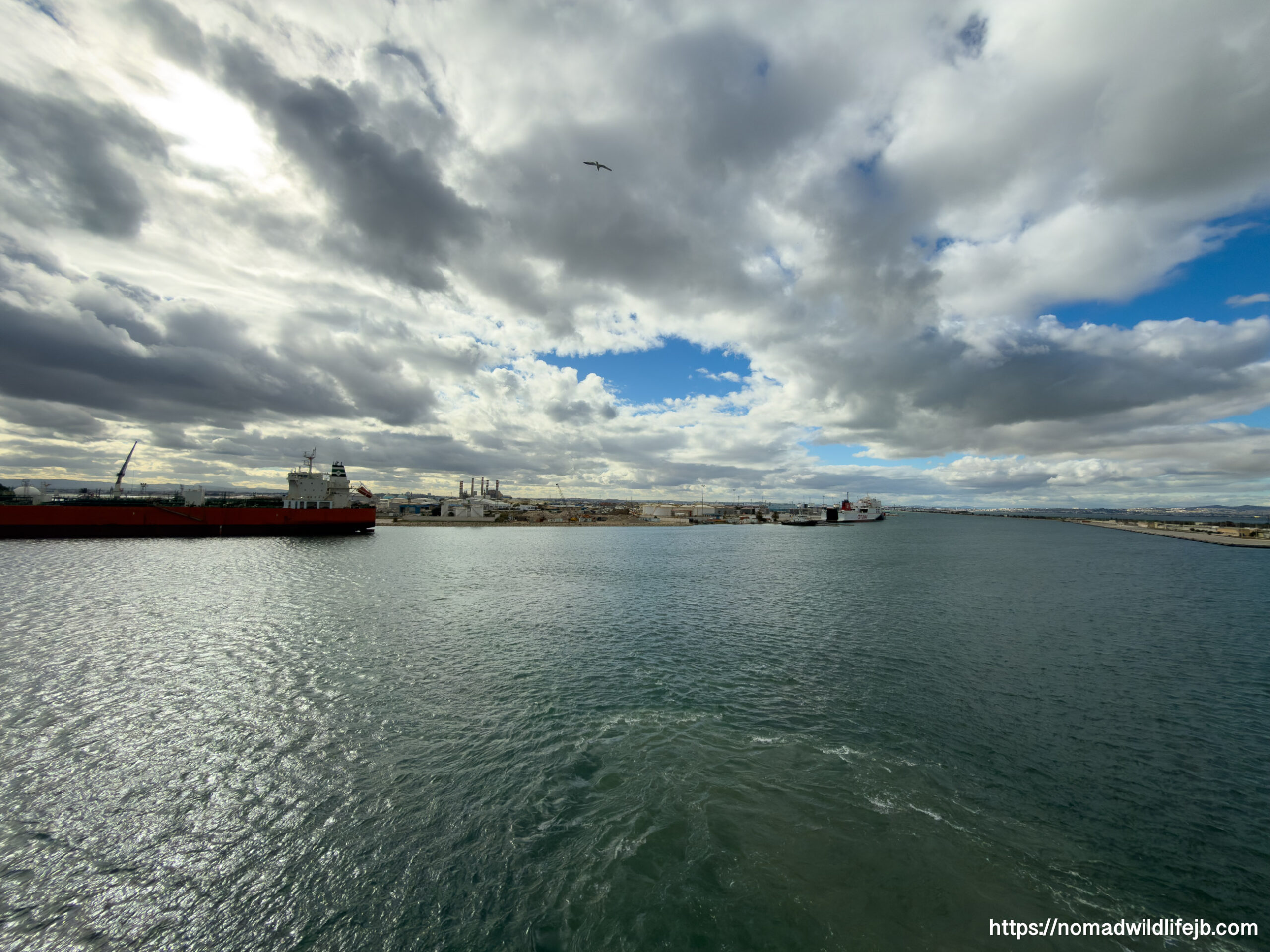 Industrial harbor with cargo ships and port infrastructure under a dramatic, cloud-filled sky, viewed from the water.