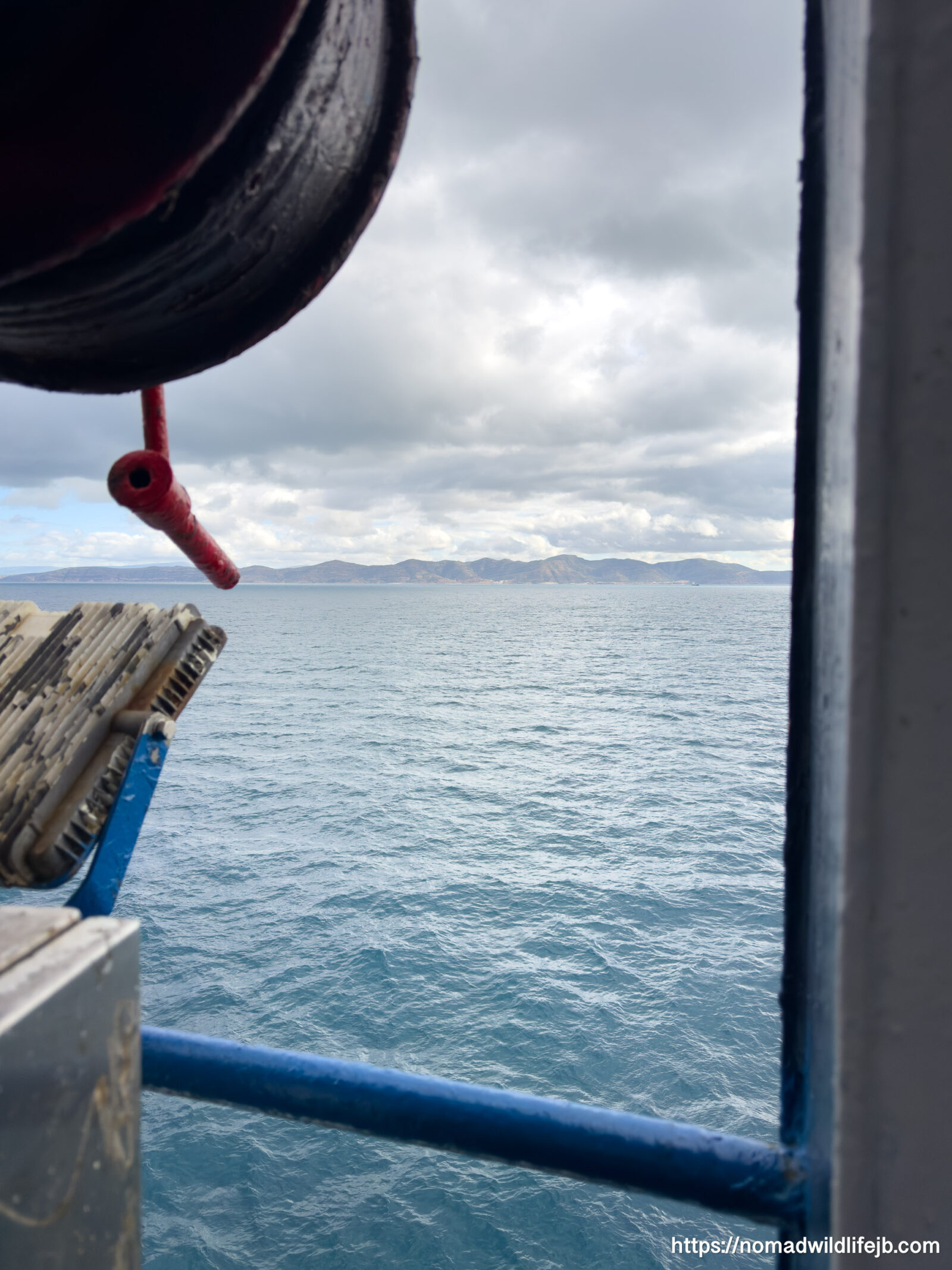 View of the Mediterranean Sea from the side of a ferry, framed by ship railings and equipment, with distant land visible on the horizon.