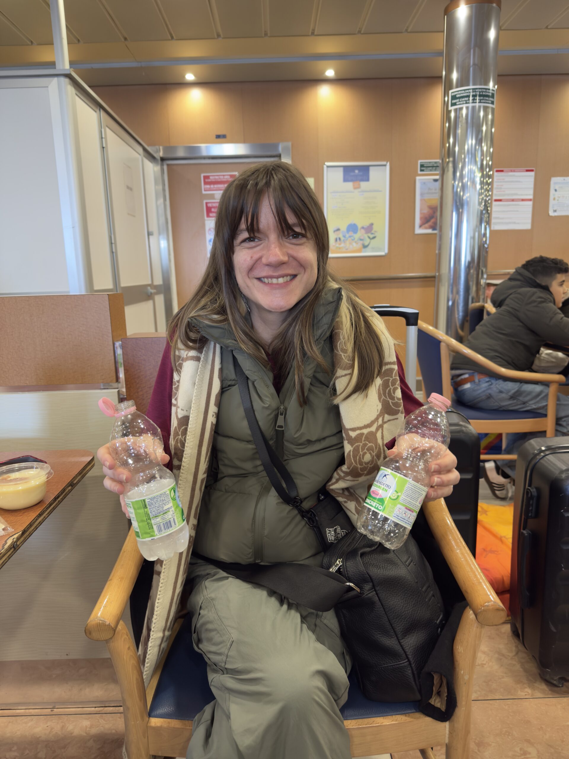 Woman seated inside a ferry passenger area, smiling while holding water bottles, with luggage and seating visible around her.