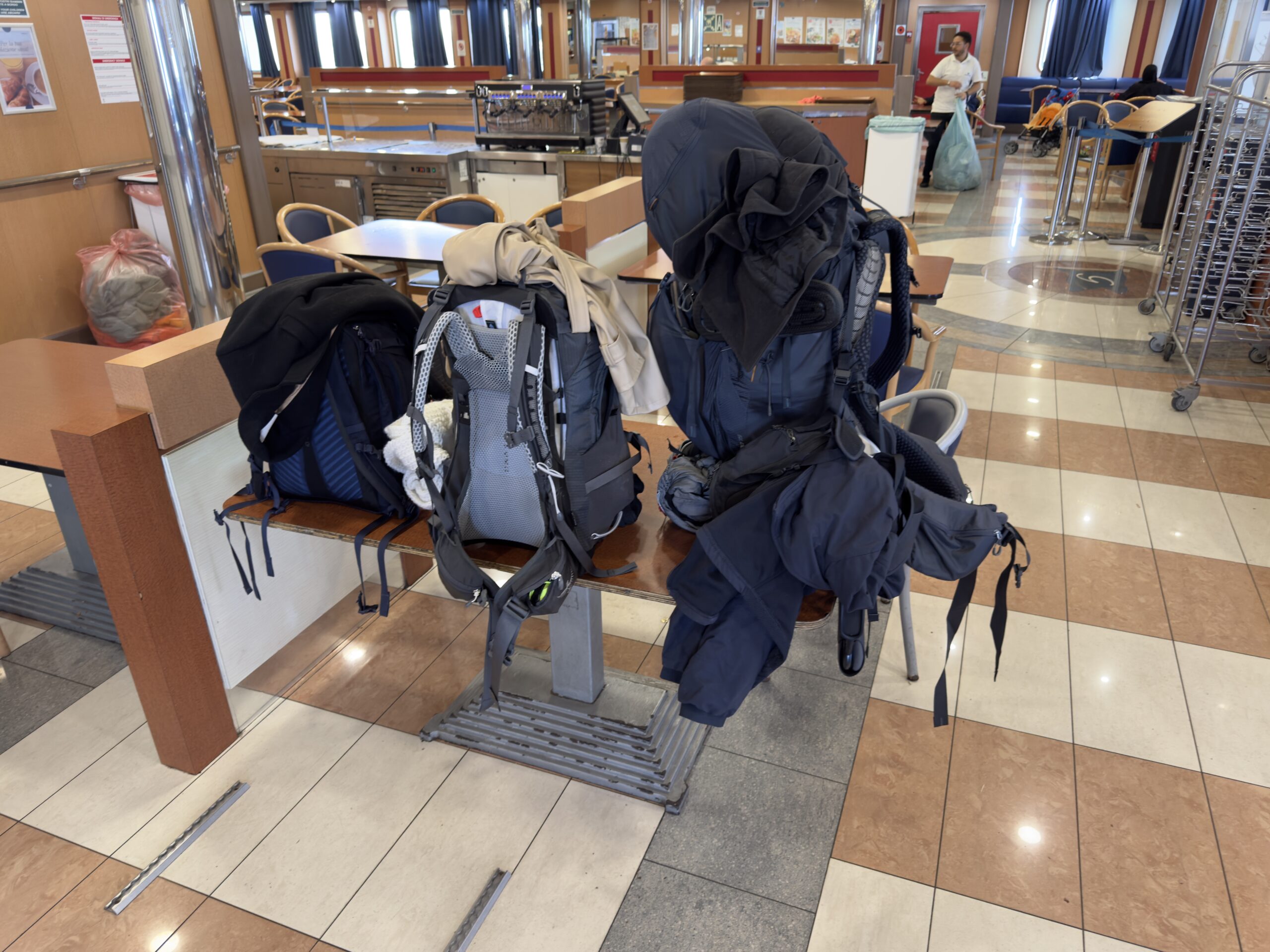 Two large backpacks resting on a bench inside a ferry passenger area, with clothing and gear bundled on top, surrounded by tables and seating.