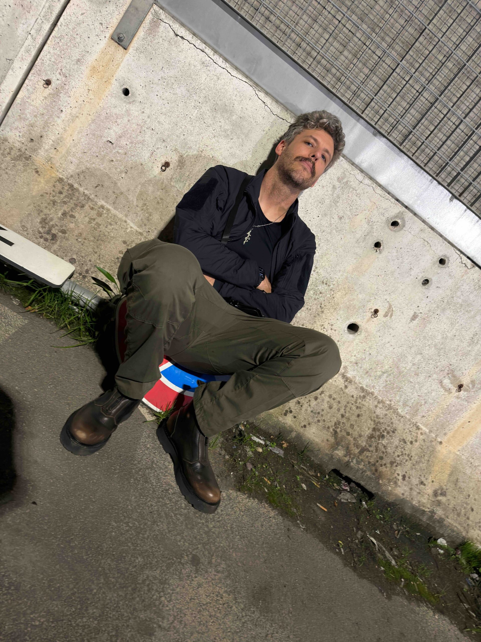 Man seated against a concrete wall in a port area, resting on a temporary seat while waiting, wearing travel clothing and boots.