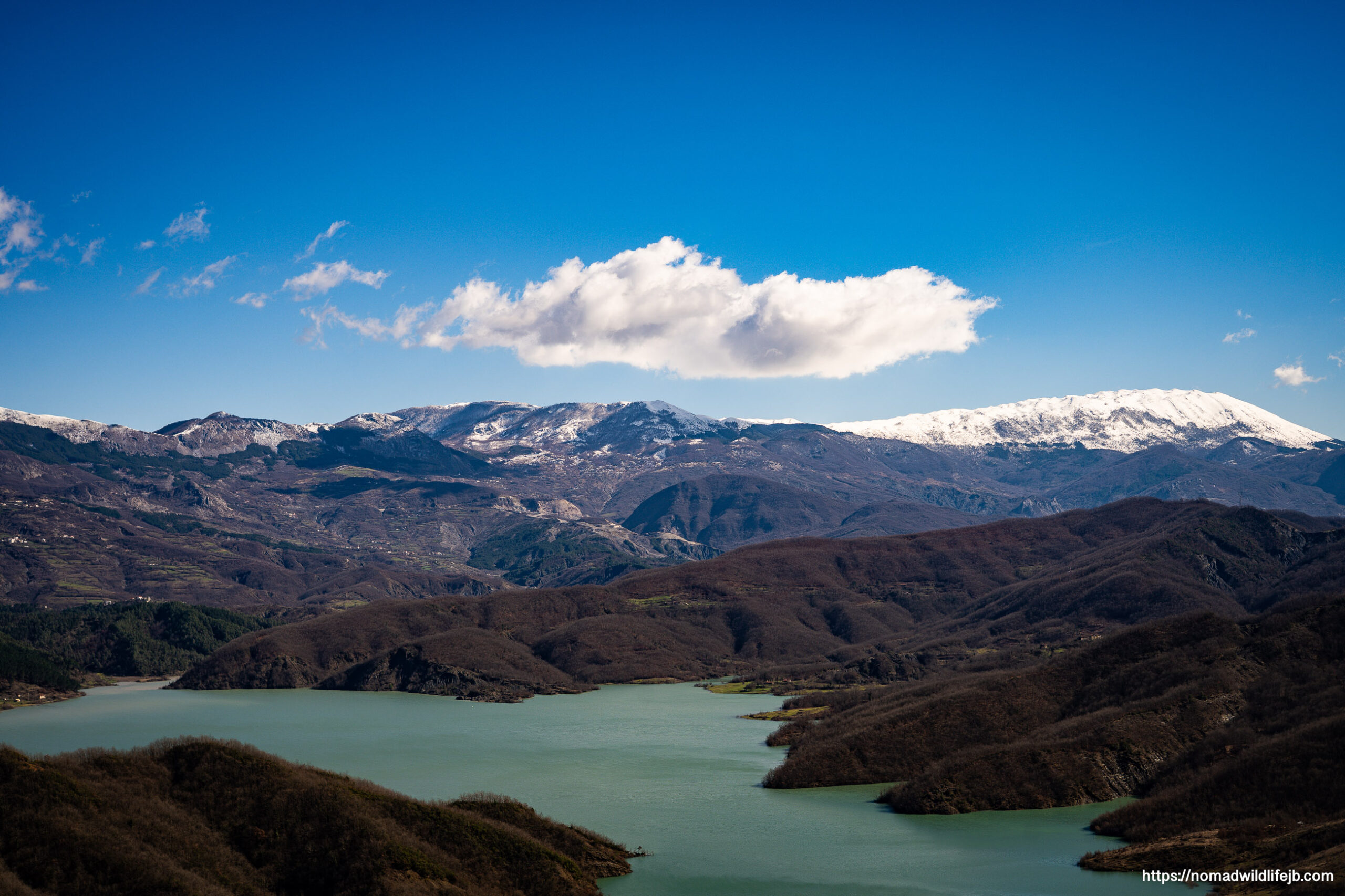 Wide panoramic view of Lake Bovilla’s turquoise water winding through dark winter hills beneath a vivid blue sky, with snowcapped mountains on the horizon.