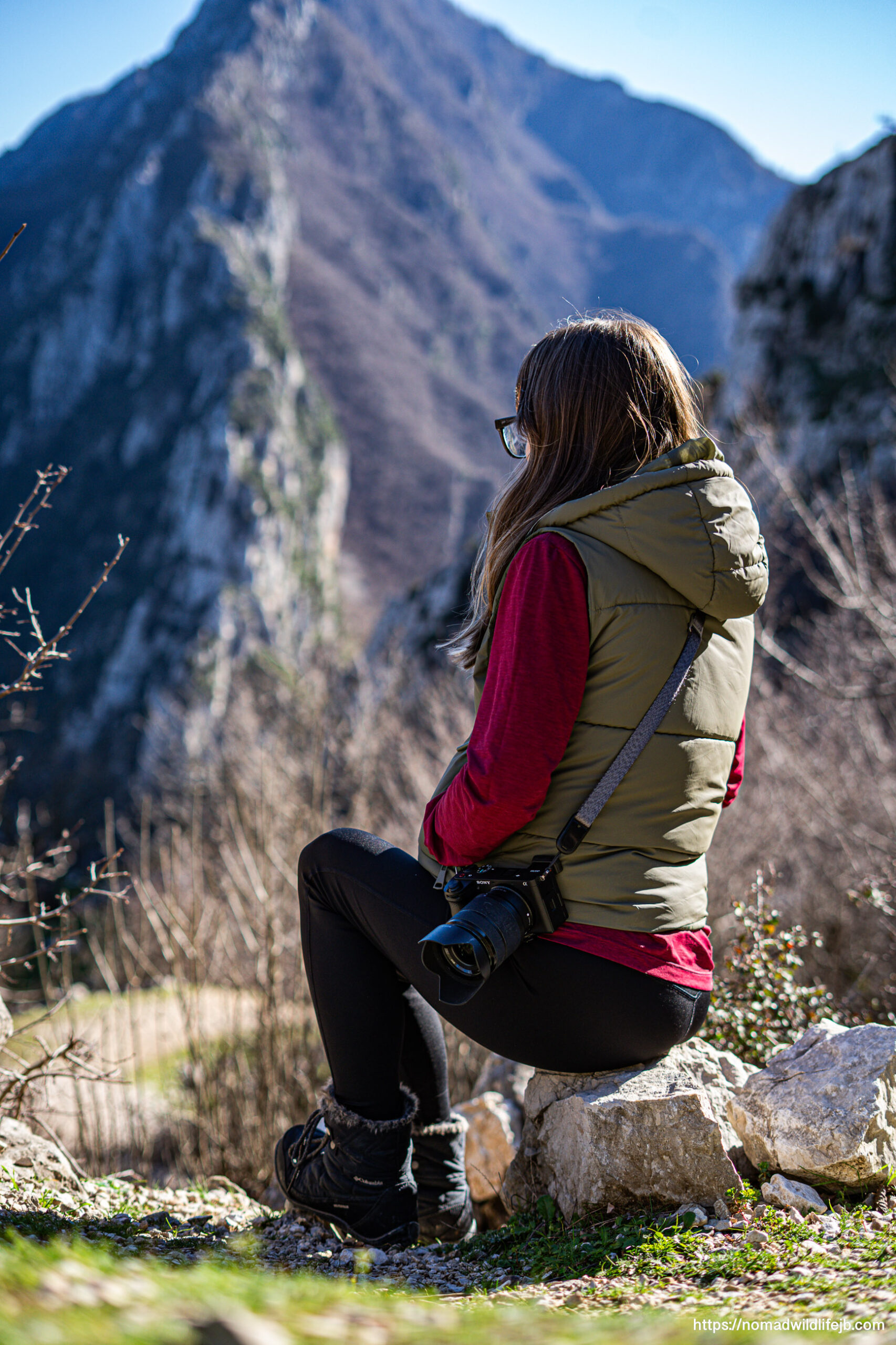 Jess sits on a rock with a camera at her side, looking out toward steep mountains in clear daylight.