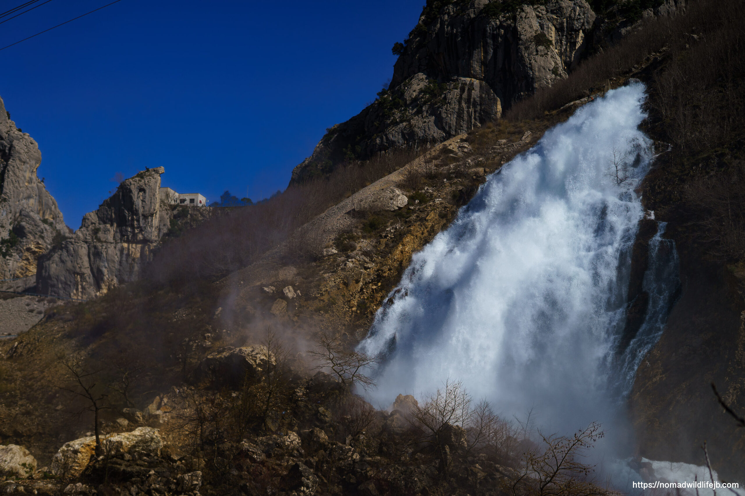 Powerful seasonal waterfall surging down a rocky mountainside near Lake Bovilla under a deep blue sky, with mist rising at the base.