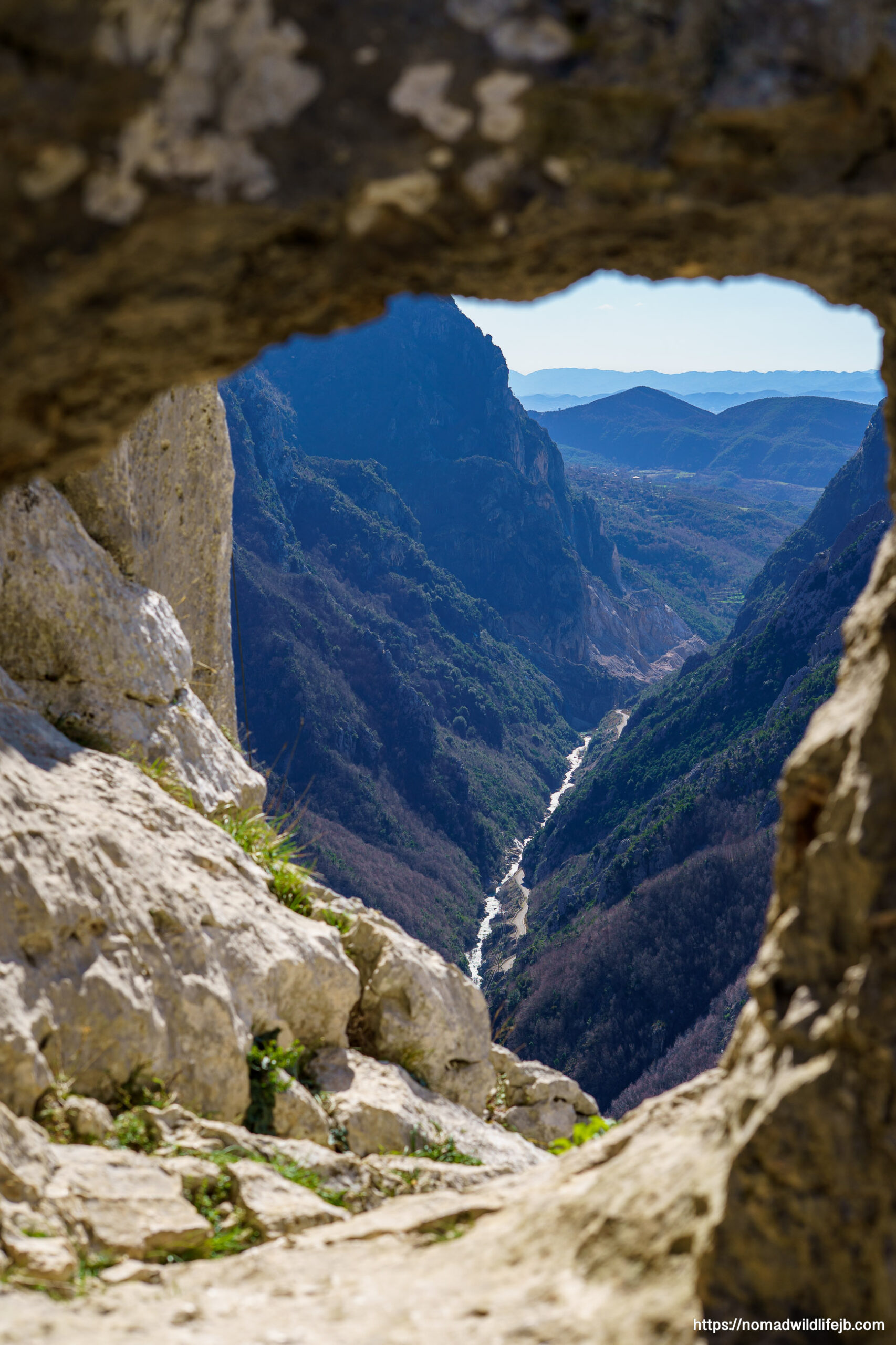 A dramatic valley view framed by rough rock like a natural window, looking down to a narrow river winding between steep mountains.