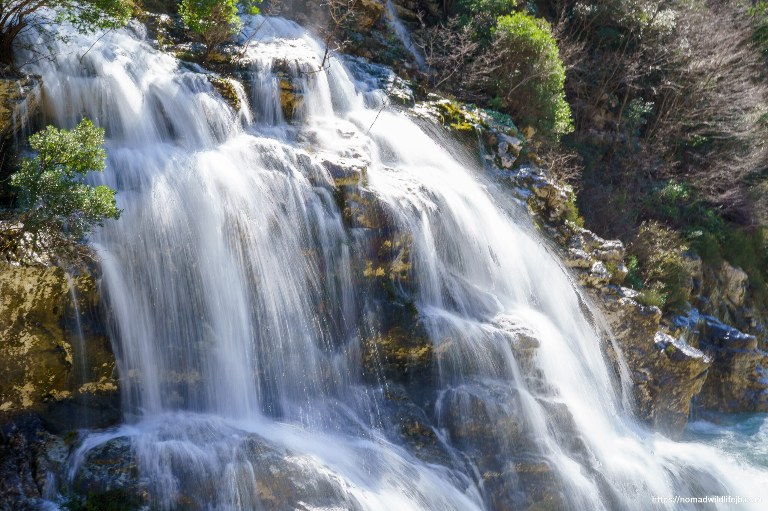 A broad waterfall cascades over layered rock in bright daylight, rendered silky by a slow shutter, with green shrubs framing the scene.