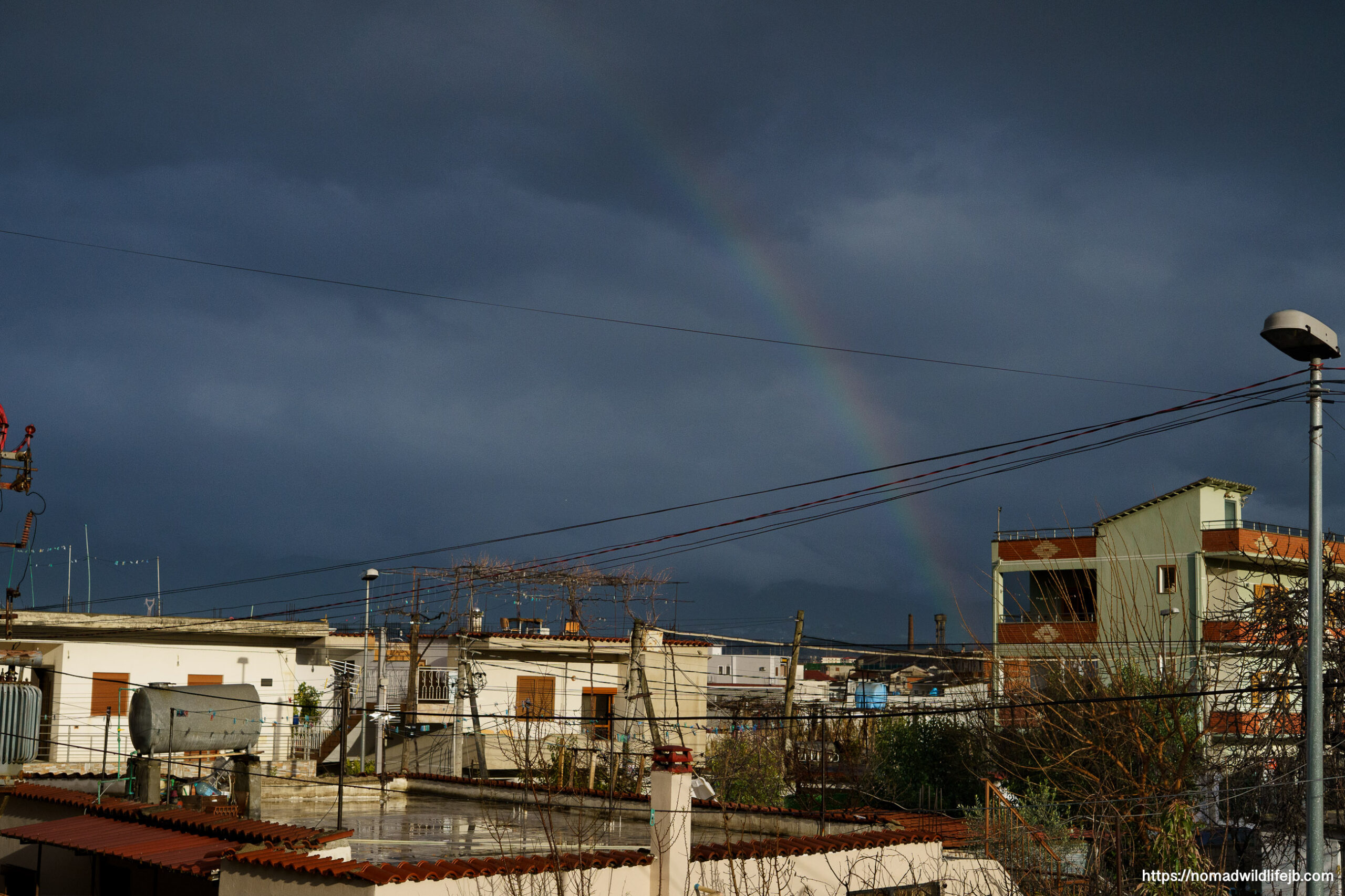 Rainbow over rooftops in Tirana, Albania beneath dark storm clouds.