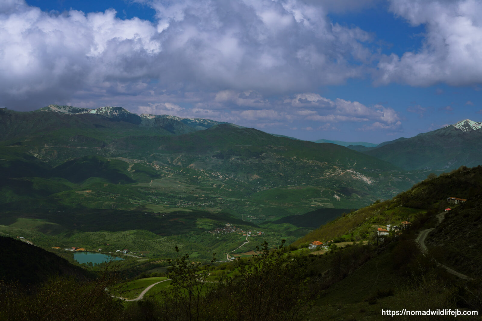 Mountain valley landscape viewed from above in Albania, with winding roads, scattered villages, and dramatic clouds over green hills.