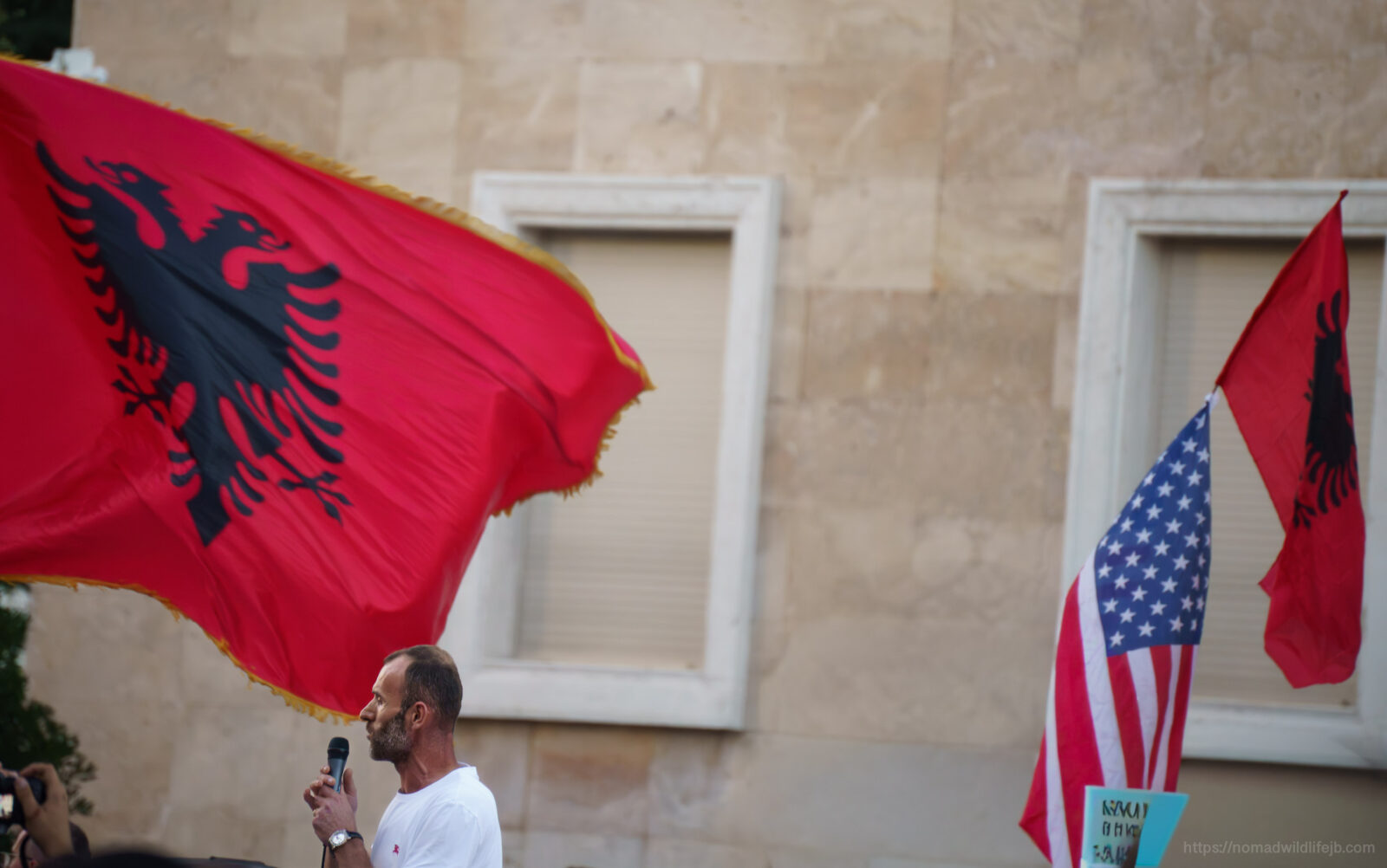 Albanian and American flags fly above a speaker addressing a protest crowd in Tirana.