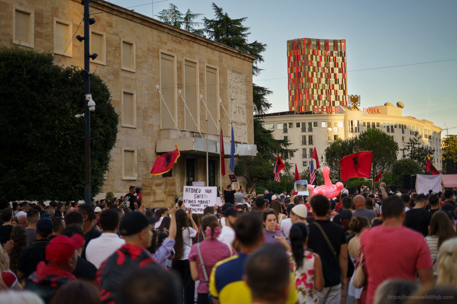 A crowd in Tirana holds Albanian and American flags around a pink flamingo float during a protest.